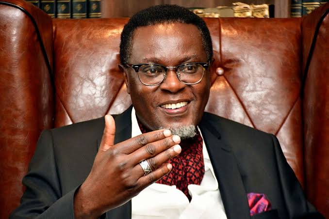 Mutahi Ngunyi seated in a leather chair in front of a bookshelf, wearing a black suit and a red cravat, with one hand raised